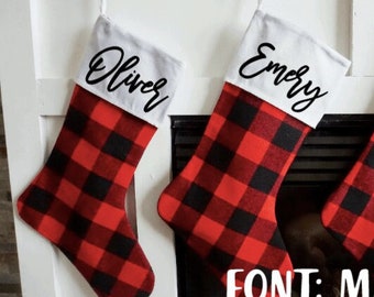 May include: Three red and black buffalo plaid Christmas stockings with white cuffs. The stockings are personalized with the names "Oliver", "Emery", and "Ryan" in a black script font called "Madina".