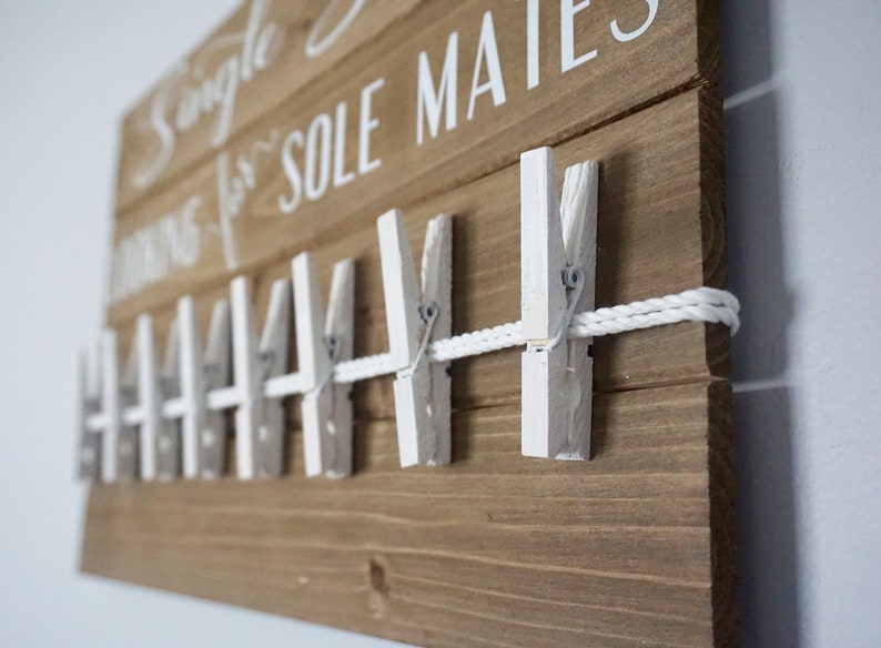 May include: A wooden sign with the text "Single looking for SOLE MATES" in white. A row of white clothespins is attached to a white rope. The wooden board is stained a medium brown colour.