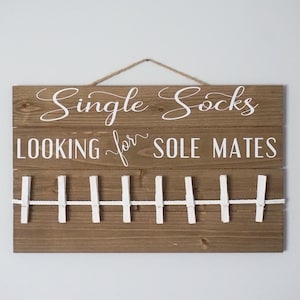 May include: A wooden sign with the words "Single Socks LOOKING for SOLE MATES" in white script. It features a clothesline with white pegs. A jute rope is used for hanging the sign.