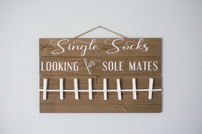 May include: A wooden sign with the words "Single Socks LOOKING for SOLE MATES" in white script. It features a clothesline with white pegs. A jute rope is used for hanging the sign.