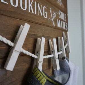 May include: Wooden sign with the text "LOOKING for SOLE MATES" in white lettering. White painted clothes pegs hold a rope and socks. The sign is made of brown wood.