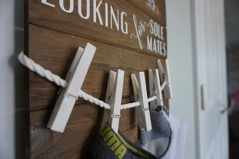 May include: Wooden sign with the text "LOOKING for SOLE MATES" in white lettering. White painted clothes pegs hold a rope and socks. The sign is made of brown wood.