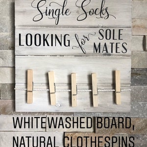 May include: Whitewashed wooden sign with the text "Single Socks LOOKING for SOLE MATES." Features a clothesline with five natural clothespins. The sign is mounted on a stone-like wall.