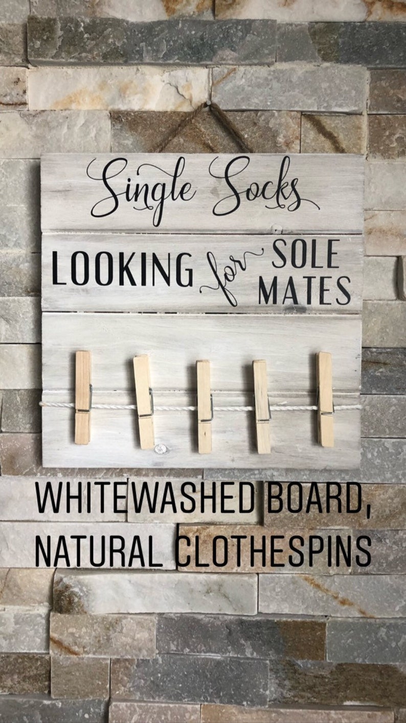 May include: Whitewashed wooden sign with the text "Single Socks LOOKING for SOLE MATES." Features a clothesline with five natural clothespins. The sign is mounted on a stone-like wall.