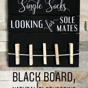 May include: Black wooden sign with white text reading "Single Socks LOOKING for SOLE MATES". It features a row of natural wooden pegs and a string for hanging. The sign is mounted on a stone wall.