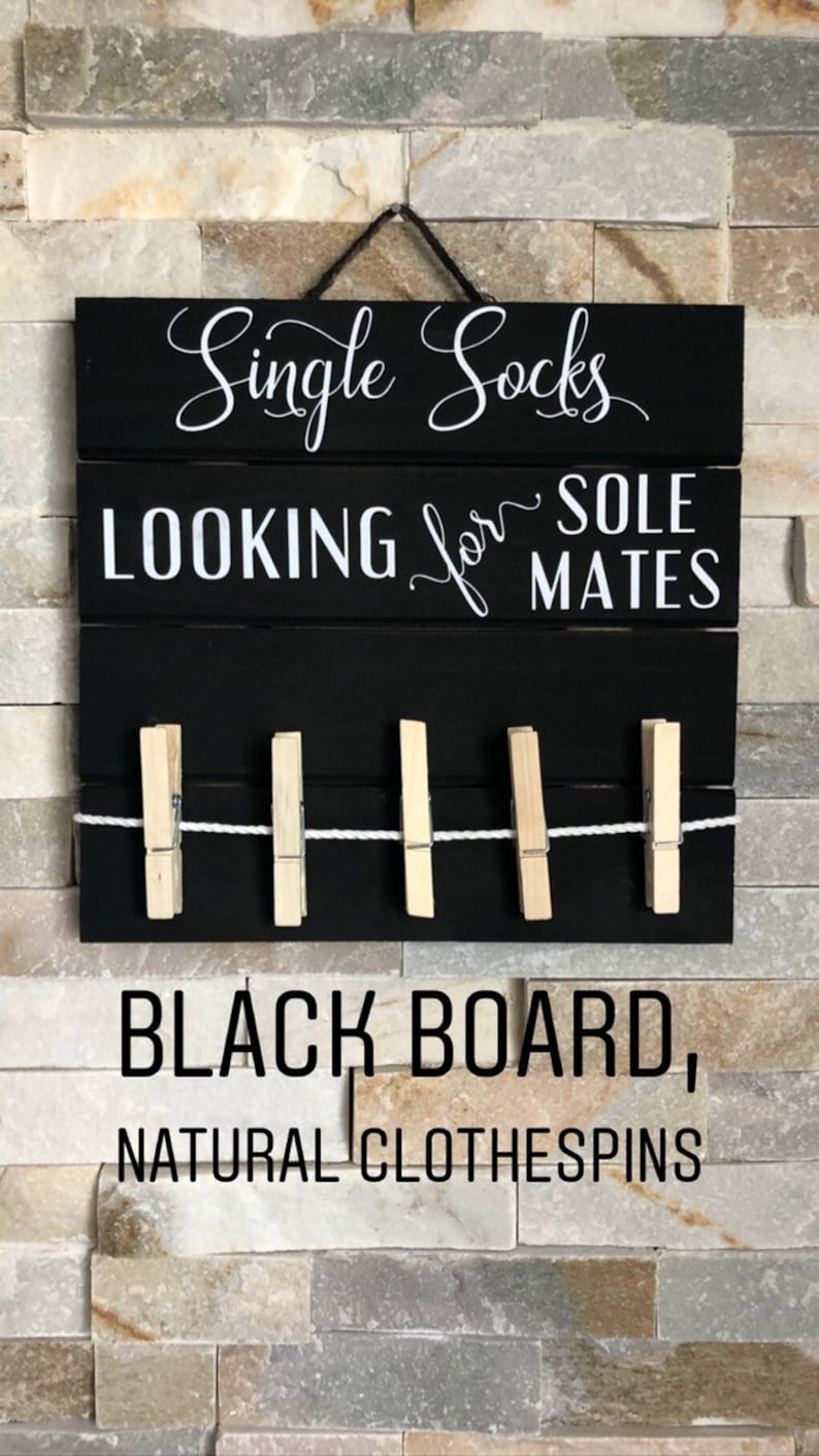 May include: Black wooden sign with white text reading "Single Socks LOOKING for SOLE MATES". It features a row of natural wooden pegs and a string for hanging. The sign is mounted on a stone wall.
