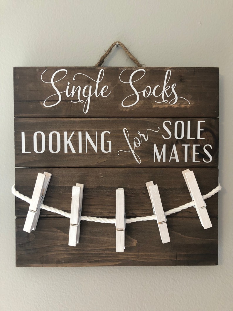 May include: A brown wooden sign with the text "Single Socks LOOKING for SOLE MATES" in white script. A white rope is strung across the bottom, held by five white clothes pegs. The sign has a rope for hanging.