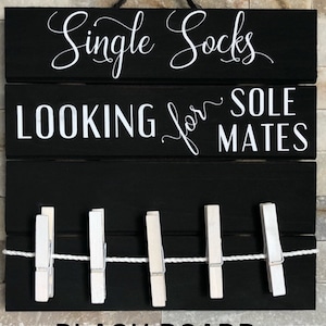 May include: Black wooden sign with white text: "Single Socks LOOKING for SOLE MATES." Features a clothesline with five white pegs. The sign is suspended against a brick wall.