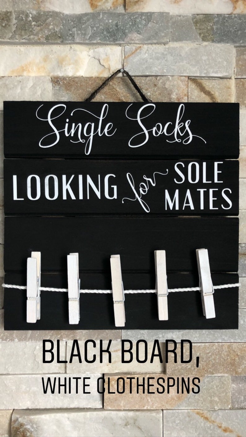 May include: Black wooden sign with white text: "Single Socks LOOKING for SOLE MATES." Features a clothesline with five white pegs. The sign is suspended against a brick wall.