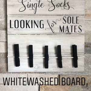 May include: A whitewashed wooden sign with the text "Single Socks LOOKING for SOLE MATES". The sign features five black clothes pegs attached with a white rope. The background is a stone wall.
