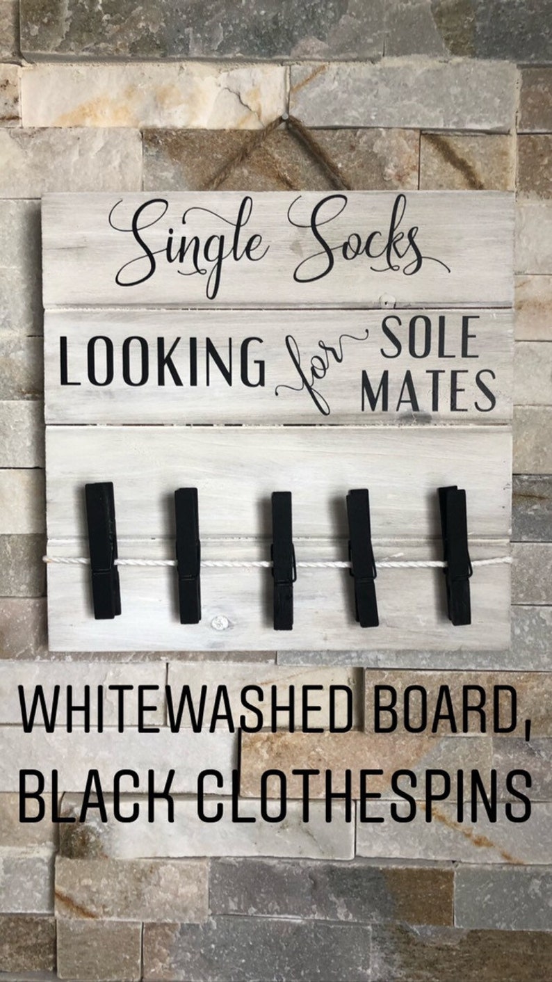 May include: A whitewashed wooden sign with the text "Single Socks LOOKING for SOLE MATES". The sign features five black clothes pegs attached with a white rope. The background is a stone wall.