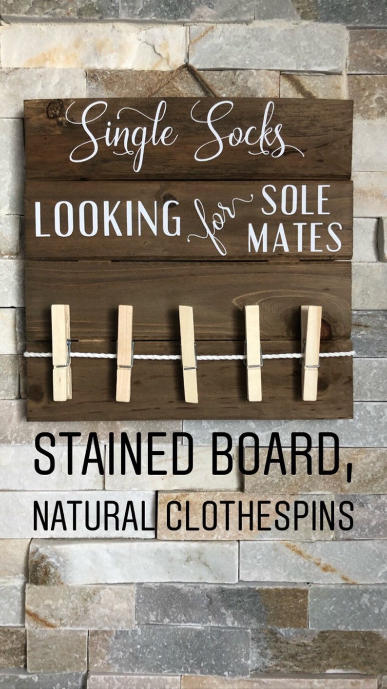 May include: A dark stained wooden sign with white lettering reading "Single Socks LOOKING for SOLE MATES". It features a clothesline with five natural wooden pegs. The sign also says "STAINED BOARD, NATURAL CLOTHESPINS".