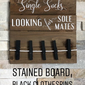 May include: A wooden sign with the text "Single Socks LOOKING for SOLE MATES". It features black clothespins and a white string. The bottom of the sign reads "STAINED BOARD, BLACK CLOTHESPINS".