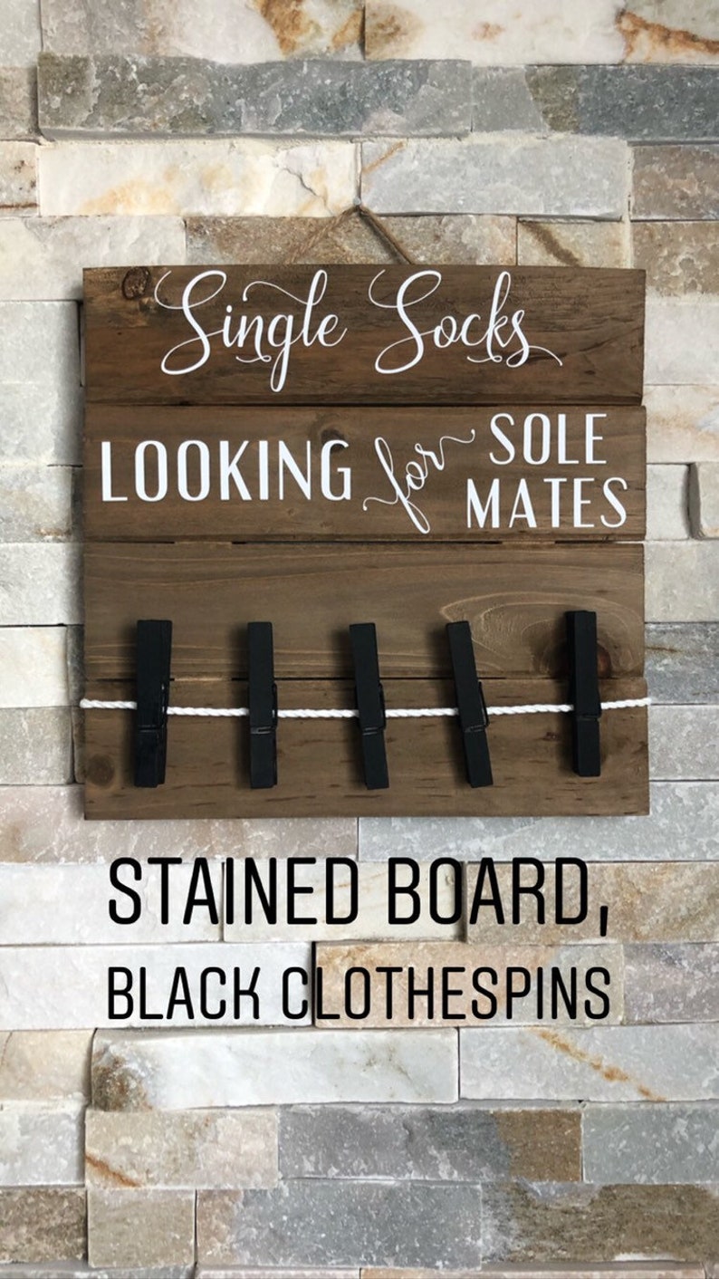 May include: A wooden sign with the text "Single Socks LOOKING for SOLE MATES". It features black clothespins and a white string. The bottom of the sign reads "STAINED BOARD, BLACK CLOTHESPINS".