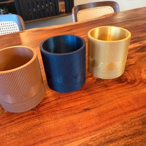 May include: Three cylindrical planters featuring a vertical ribbed texture and a repeating arched pattern around the base. The planters are finished in matte terracotta, dark navy blue, and metallic gold colors, each with an open top design.
