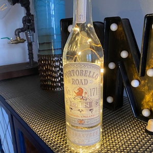 May include: A clear glass bottle with a gold label that reads "Portobello Road No. 171 London Dry Gin" and "London, England". The bottle is illuminated with fairy lights.
