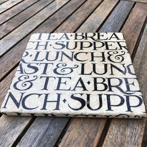 May include: A square, off-white stone tile featuring black, serif-style typography. The text repeats words related to meals, including TEA, BREAKFAST, LUNCH, and SUPPER, separated by small diamond-shaped symbols in a repeating, stacked pattern.