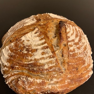 K&ouml;nnte beinhalten: Ein frisch gebackenes Sauerteigbrot mit einer knusprigen Kruste und einer leichten Mehlbest&auml;ubung. Das Brot hat ein rustikales Aussehen mit einem tiefen Einschnittmuster.