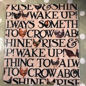 May include: Square ceramic tile with a beige background and black text that reads "RISE & SHINE! WAKE UP! ALWAYS SOMETHING TO CROW ABOUT." The tile features illustrations of chickens.
