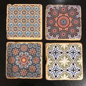 May include: Set of four square coasters featuring intricate geometric patterns in shades of blue, green, yellow, red, and black. The coasters are made from stone and have a rustic, handmade appearance.