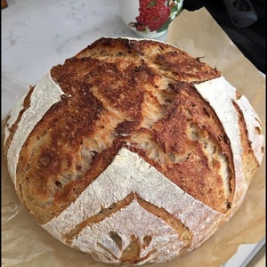 K&ouml;nnte beinhalten: Ein frisch gebackenes Sauerteigbrot mit einer knusprigen braunen Kruste und einem wei&szlig;en Inneren. Das Brot hat ein dekoratives Muster auf der Oberseite.