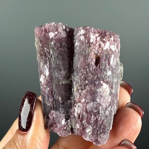 May include: A natural mineral specimen in shades of purple. The crystalline structure has white inclusions. The mineral is held in a hand with dark red nail polish.