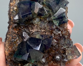 Könnte beinhalten: Ein Cluster aus dunkelgrünen Fluoritkristallen mit einem metallischen Glanz, eingebettet in eine Matrix aus braunem Gestein. Die Kristalle sind scharf und gut definiert, mit einer Vielzahl von Größen und Formen.