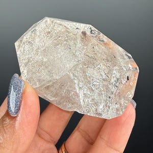 Puede incluir: Un cristal de cuarzo transparente con una forma geométrica multifacética. El cristal se sostiene en una mano, mostrando su cualidad translúcida e inclusiones internas. El cuarzo tiene una apariencia natural y en bruto, con sutiles variaciones de color.