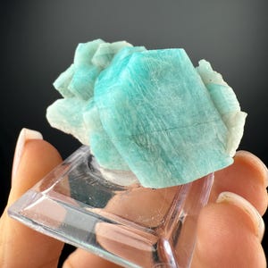 Puede incluir: Primer plano de un grupo de cristales de amazonita azul claro. El cristal está montado sobre un expositor de plástico transparente y cuadrado. La amazonita tiene una estructura cristalina y un color azul verdoso pálido.