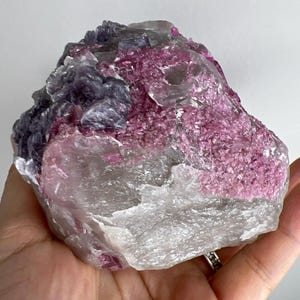 Op de afbeelding: Een grote, ruwe, roze en paarse kristalkluster met een heldere, doorschijnende basis. De kristal heeft een ruwe, natuurlijke textuur.