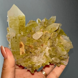 Puede incluir: Un cúmulo de cristales de cuarzo transparentes con inclusiones verdes. Los cristales están agrupados en una formación natural, con algunos de los cristales apuntando hacia arriba.