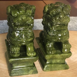 Może przedstawiać: Para zielonych jadeitowych posągów foo dog, każda stojąca na kwadratowej podstawie. Posągi foo dog są misterne rzeźbione z precyzyjnymi detalami i błyszczącym wykończeniem.