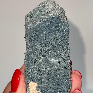 May include: A dark blue crystal cluster with a rough, textured surface. The crystal is held in a hand with red nail polish. The base of the crystal has a beige, rough texture.