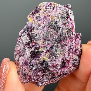 May include: A raw, unpolished mineral specimen featuring a deep magenta and dark purple crystalline structure with scattered flecks of yellow and translucent white inclusions throughout the rough, jagged, and uneven surface of the stone.
