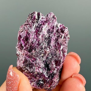 May include: A raw, unpolished mineral specimen featuring a deep magenta and dark purple colour palette. The stone has a jagged, crystalline texture with reflective, metallic-looking surfaces and irregular edges throughout its entire structure.
