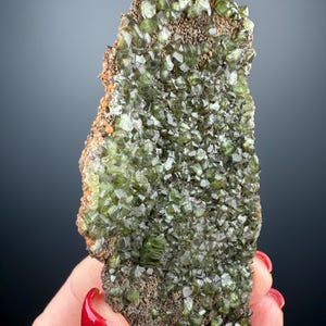 May include: A cluster of green, glassy crystals on a dark gray matrix. The crystals are sharply formed and have a slightly metallic sheen.