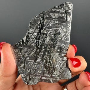 May include: A polished slice of iron meteorite featuring a distinct Widmanstätten pattern of intersecting metallic lines and dark, textured inclusions. The surface displays a reflective, silver-toned finish with geometric crystalline structures.