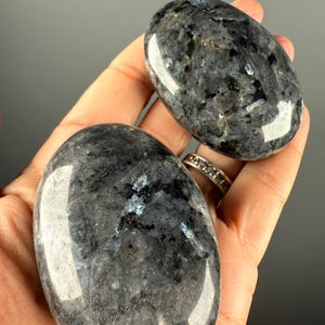 Pode incluir: Duas pedras de palma de labradorite polida. As pedras têm formato oval com uma mistura de tons cinza, preto e azul iridescente. As pedras são lisas e refletem a luz, mostrando seus padrões naturais. Ideais para meditação.