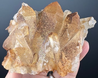 May include: A cluster of quartz crystals with a golden-brown hue and speckled patterns. The crystals are sharply pointed and arranged in a layered formation, showcasing natural geological beauty. The quartz is translucent, allowing light to pass through.