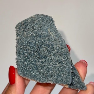 May include: A piece of blue-grey mineral with a rough, textured surface. The mineral has a crystalline appearance and is held in a hand with red nail polish.