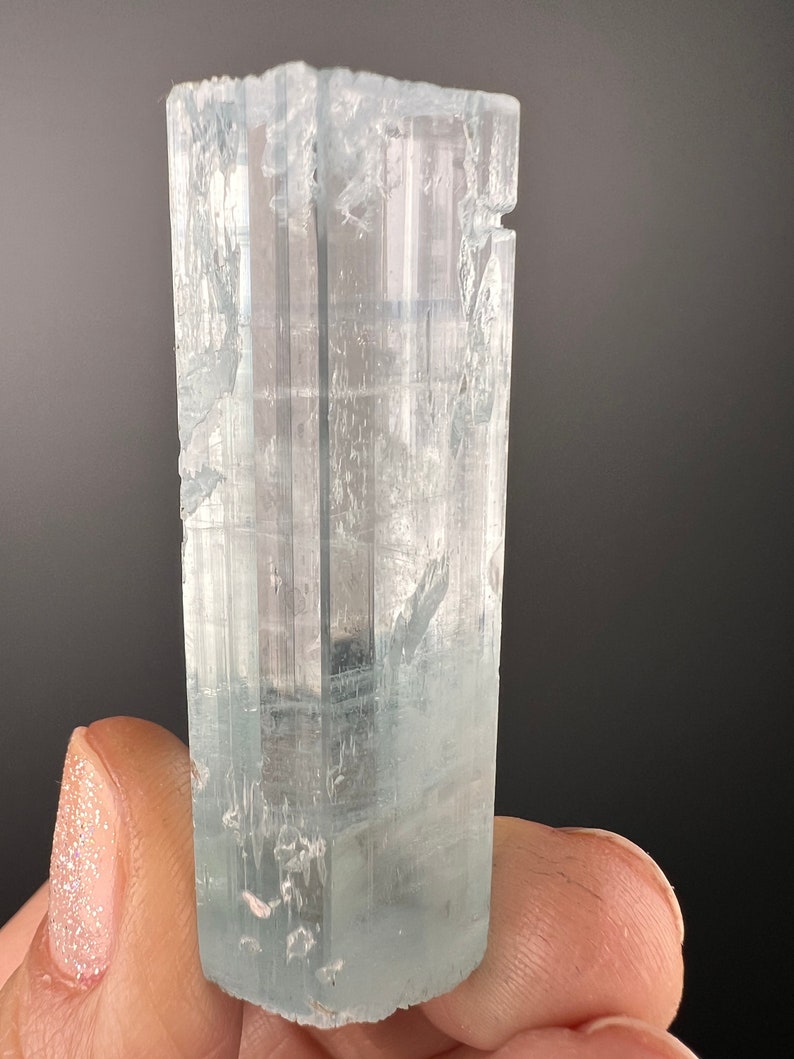 May include: A light blue aquamarine crystal with a faceted, rectangular shape. The crystal is translucent and has a slight green hue.