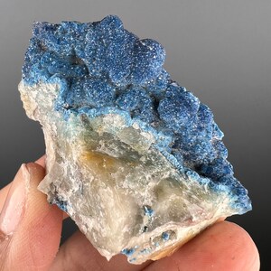 Op de afbeelding: Een close-up van een mineraal specimen met een levendige blauwe, kristallijne formatie bovenop een witte en bruine basis. De blauwe kristallen hebben een textuur, bobbelig uiterlijk. Het specimen wordt in de hand gehouden.