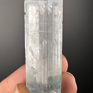 May include: A large, clear aquamarine crystal with a light blue hue. The crystal is faceted and has a rectangular shape. It is sitting on a small, white rock.
