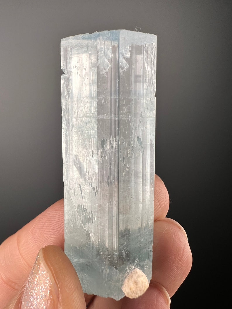 May include: A large, clear aquamarine crystal with a light blue hue. The crystal is faceted and has a rectangular shape. It is sitting on a small, white rock.