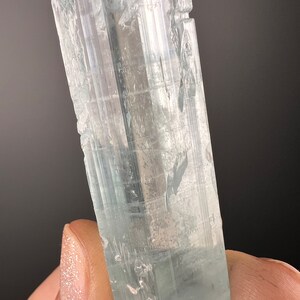 May include: A clear, light blue aquamarine crystal with striations and a slightly cloudy appearance. The crystal is approximately 2 inches long and has a hexagonal shape.