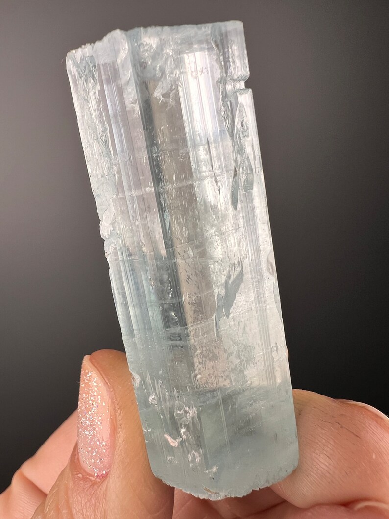 May include: A clear, light blue aquamarine crystal with striations and a slightly cloudy appearance. The crystal is approximately 2 inches long and has a hexagonal shape.