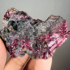 May include: A rough, gray rock with bright pink and red crystals embedded in it. The crystals are clustered together and have a metallic sheen.
