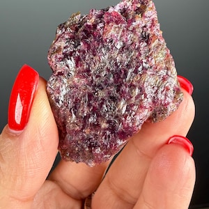 Könnte beinhalten: Ein raues, unregelmäßig geformtes Mineralexemplar mit tiefmagentafarbener Tönung, gesprenkelt mit Gold- und Silbertönen. Die kristalline Struktur ist sichtbar und erzeugt eine strukturierte Oberfläche. Das Mineral wird in einer Hand mit rotem Nagellack gehalten.