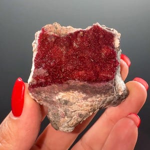 Puede incluir: Un espécimen mineral natural con una vibrante capa cristalina roja sobre una base de piedra rugosa, texturizada, gris y beige. Los cristales rojos están densamente empaquetados, creando una apariencia brillante. La forma general es irregular.