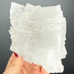 May include: A translucent, crystalline mineral specimen. The mineral is a light, off-white color and has a layered, geometric structure. The specimen is held in a hand against a dark background.